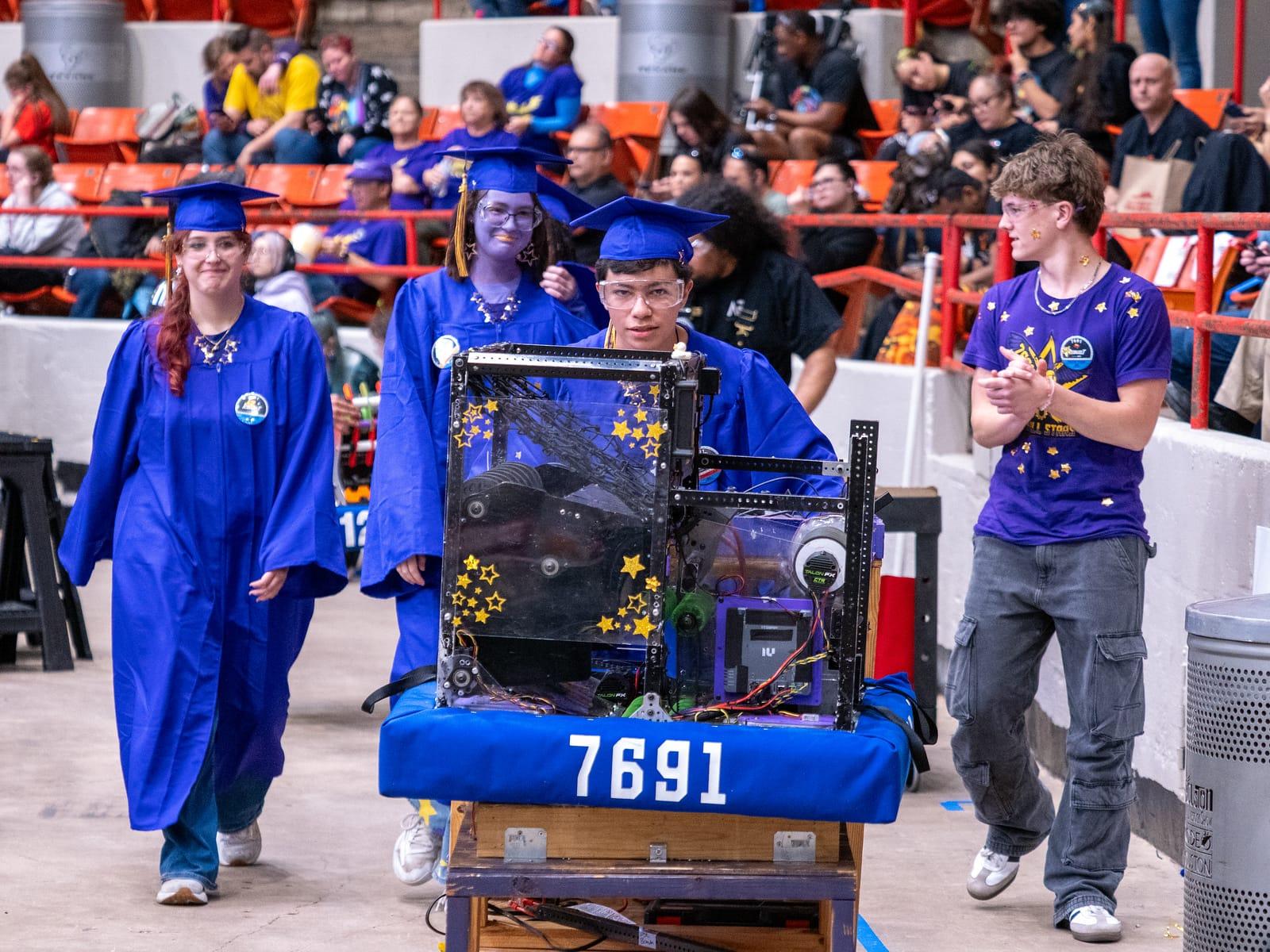Graduating seniors in caps and gowns walking their robot onto the floor at the 2026 District Championship
