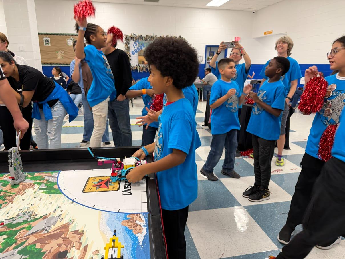 A student running their robot at the FIRST LEGO League table while teammates cheer behind him with pom-poms