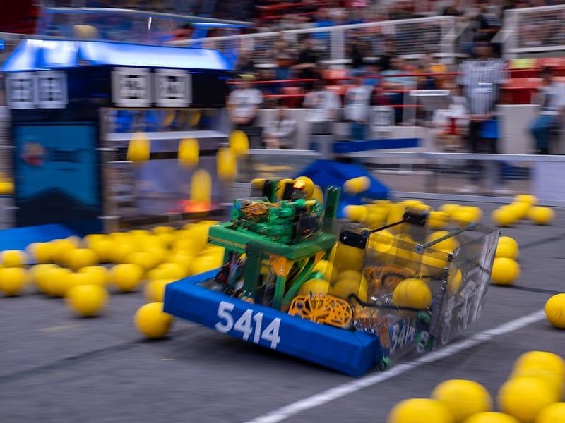A FIRST Robotics Competition robot the size of a washing machine crossing the arena floor among yellow game pieces