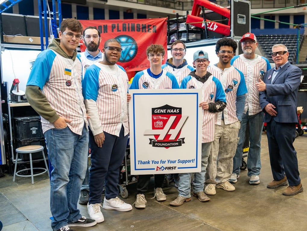 Nine students and mentors from a FIRST Robotics Competition team standing in the pits around a large board reading “Gene Haas Foundation — Thank You for Your Support!”